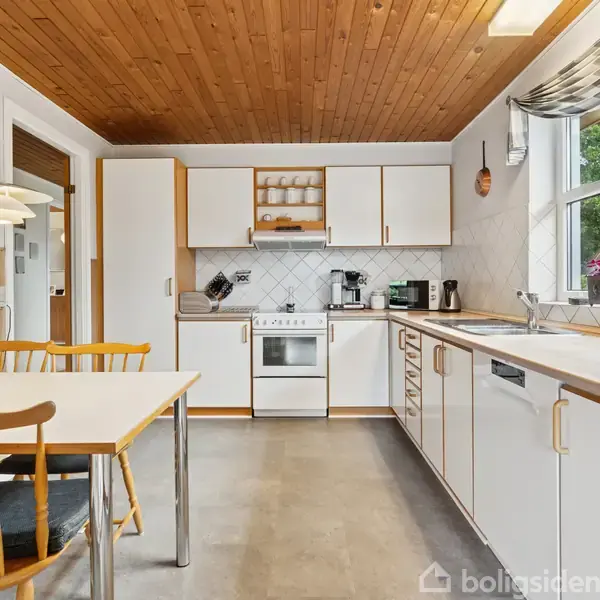 Et moderne køkken med hvide skabe og komfur i midten. Spisebord med to stole står ved siden af et vindue, der giver udsigt til en grøn have.