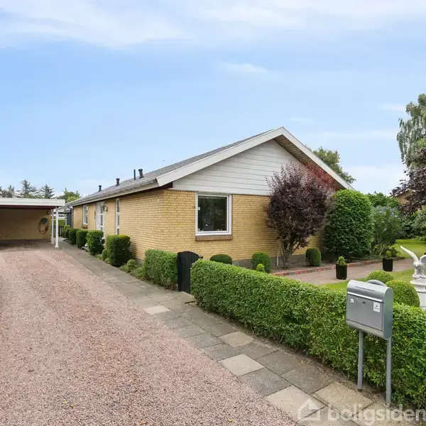 Et gult murstenshus står stille i en velplejet have med græs, buske og træer, set fra en grusbelagt indkørsel med en carport til venstre.