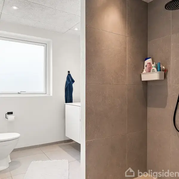 Et moderne badeværelse med hvidt toilet til venstre. Bruseniche til højre har bruser og hylder med toiletartikler. Rummet har hvide vægge, et vindue og en håndvask med håndklæde.