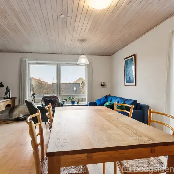 Langt spisebord med træstole står i en lys stue. Rummet har en blå sofa, skrivebord og store vinduer, som fører ud til en altan. Ingen tekst til stede.