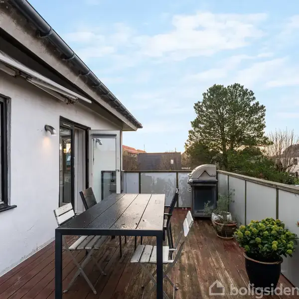 Terrasse med træbord og stole, stilleben stående ved glasrækværk. Solnedgang spejler sig i vinduet. Omgivet af huse og træer. Grill og planter står langs kanten.