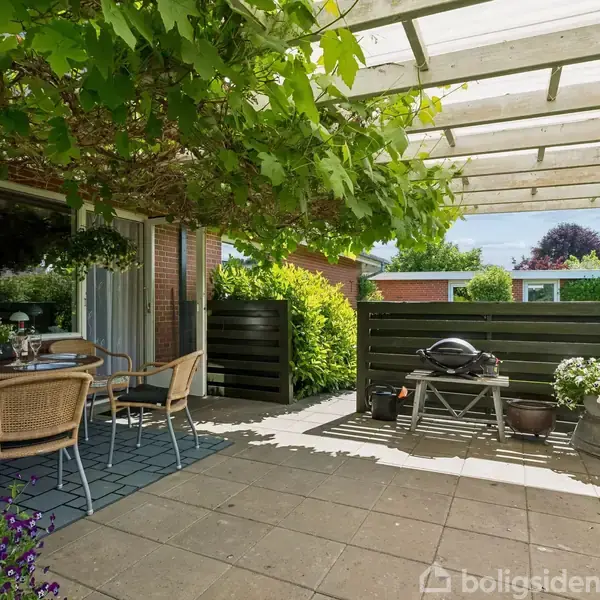 Terrasse set med flisebelagt gulv, havemøbler og grill, dækket af en pergola med grønne planter. I baggrunden ses en have med træer og buske.