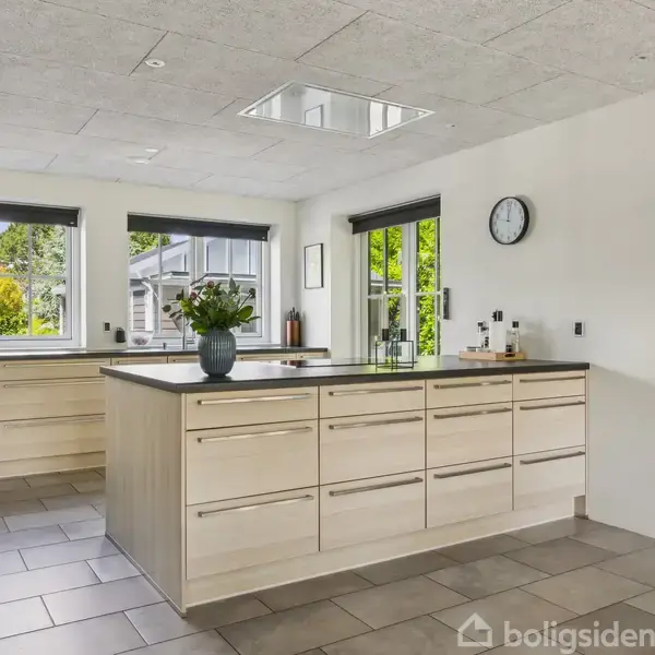 Køkkenø med vaser står i moderne køkken, omgivet af skabsmoduler og store vinduer, der indrammer en grøn udsigt. En vægur hænger på væggen, gulvet er flisebelagt.