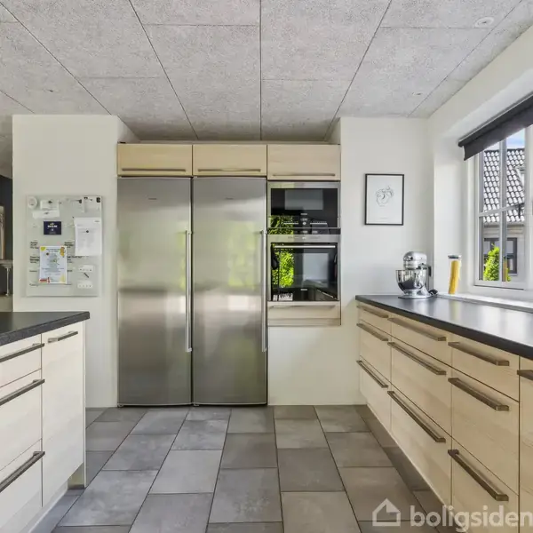 Et moderne køkken med lyse trælåger, to store stål-apparater i midten, og overskabe. Der er en stor vask under et vindue til højre, omgivet af hvide vægge.