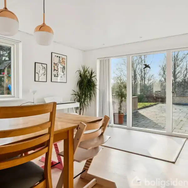 Træbord med stole står i spisestue ved store glasdøre, der åbner ud mod terrasse. Indendørs planter og billeder hænger på væggen. Sollys strømmer ind fra venstre vindue.