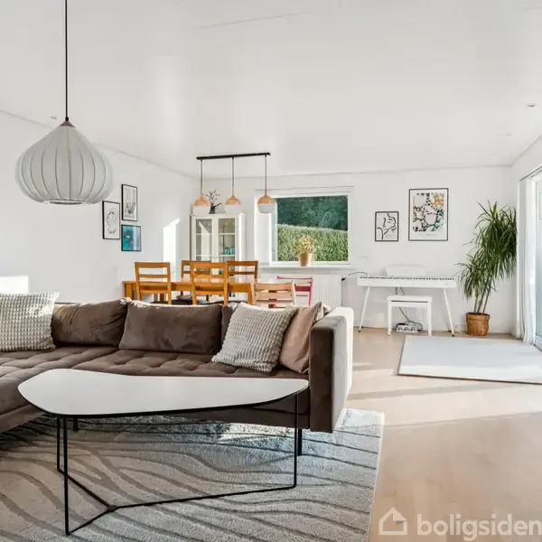 Stor hjørnesofa i brun nuance står i moderne stue med lyse vægge. Foran sofaen er et hvidt sofabord. I baggrunden ses spisebord og stol samt en stor plante nær vinduet.