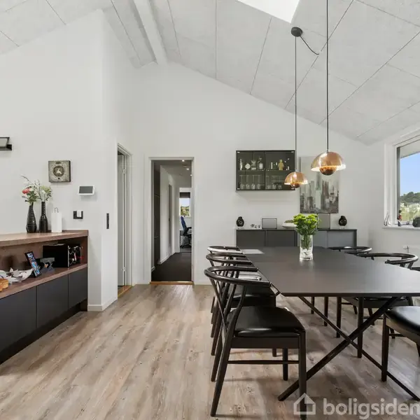 Et elegant spisebord med sorte stole står i et lyst rum med trægulv. Vaser med blomster pryder bordet. Store vinduer og loftlamper giver rummet et moderne præg.