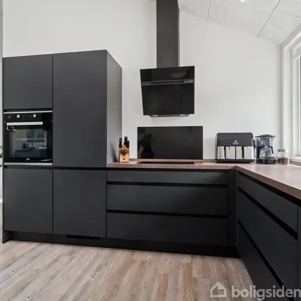 Sort køkken med moderne skabe, indbygget ovn og emhætte. Stuevæg med farverigt maleri og planter ses i baggrunden. Store vinduer til højre lader naturligt lys ind.