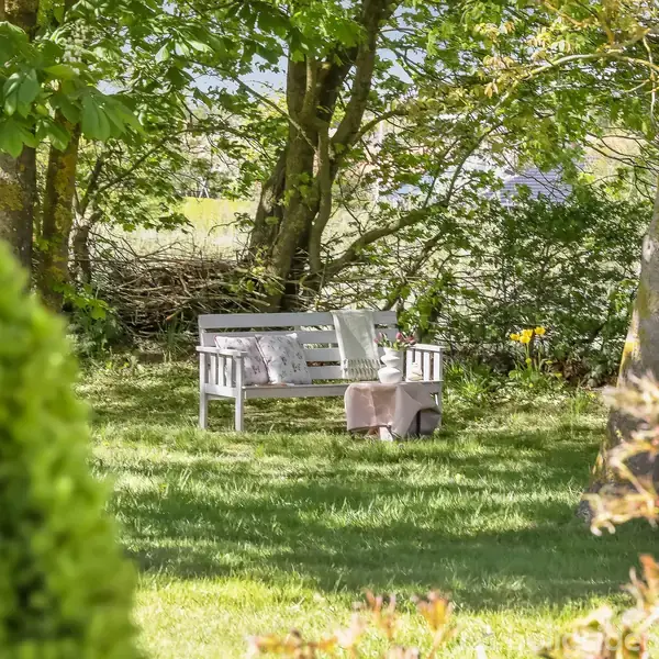En hvid bænk med puder står i en solrig have, omgivet af træer og buske, der skaber en skyggefuld og fredfyldt atmosfære.