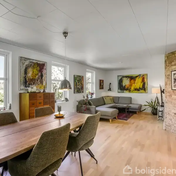 Spisebord med stole står i en stue, oplyst af hængelamper. Rummet har lyse vægge, store vinduer, kunstmalerier og en hjørnesofa foran en rå murstensvæg.