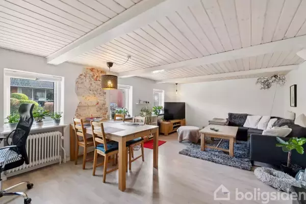 Et lyst stueområde med hvidt træloft og trægulv indeholder et spisebord med stole og en stue med sofa, sofabord og TV. Vinduer med planter tilbyder naturligt lys.