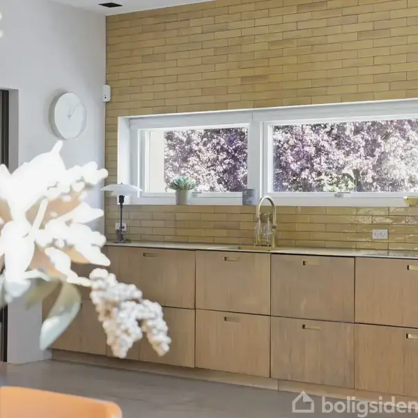 Køkkenbord med træskabe står foran en væg med gule mursten. Et vindue med udsigt til lilla træer er ovenfor, og en blomstervase står i forgrunden.
