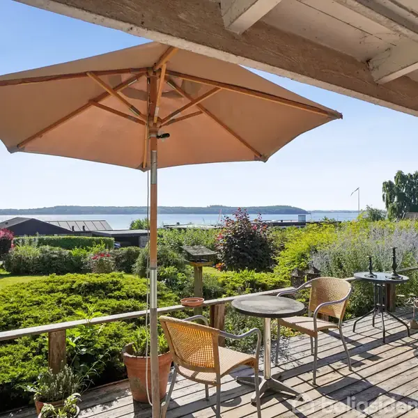 En terrasse med to stole og et bord under en stor parasol. Terrassen har udsigt over en frodig have med buske og en sø i baggrunden.