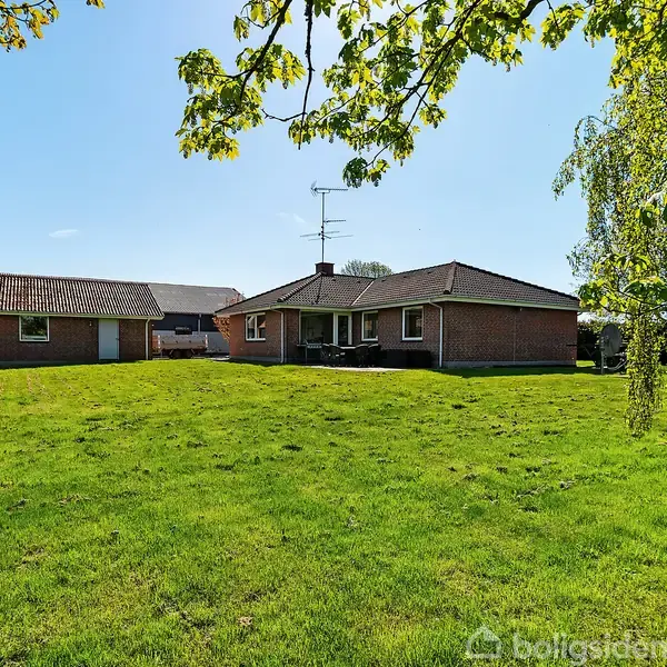 En rød murstensbungalow med fladt tag står i en grøn have med spredte træer og under en blå himmel. Der ses også en garagebygning til venstre.