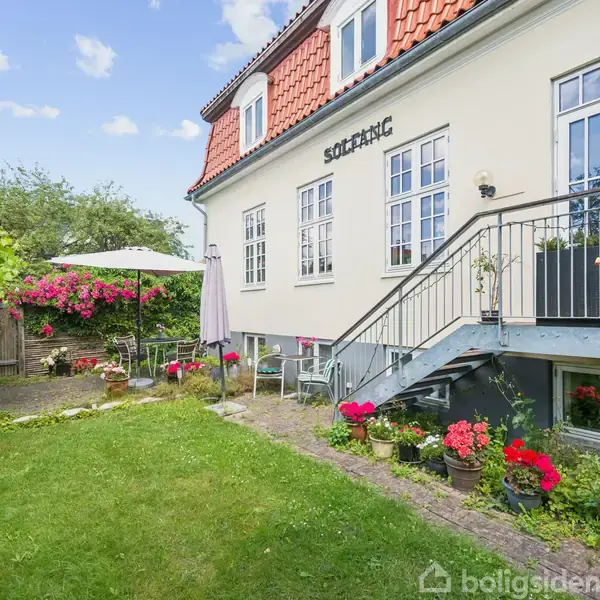 Hus med navnet "Solvang" malet på væggen, har en trappe op til en terrasse. Der er blomster i krukker langs trappen og en græsplæne i en frodig have med siddeplads.
