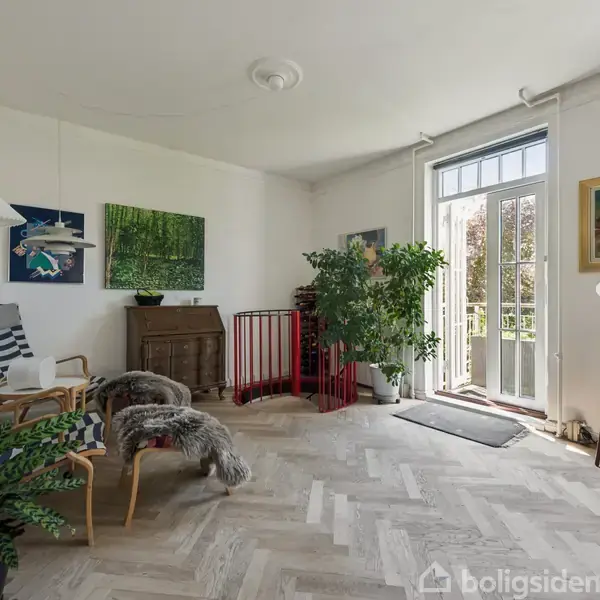 To stole med stribede hynder står ved siden af et lille bord i en stue. Store planter og kunstværker dekorerer rummet, der har åbne franske døre mod en balkon.