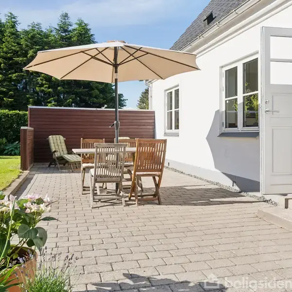 Terrassebord med stole står under en parasol på en flisebelagt terrasse ved siden af et hvidt hus. Der er græsplæne og tæt buskads i baggrunden, blomster i forgrunden.