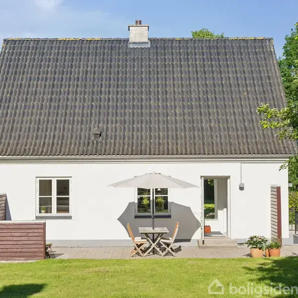 Et hvidt hus med sort tag står i en grøn have. En terrasse foran rummer et bord med parasol og stole, omgivet af træværk.