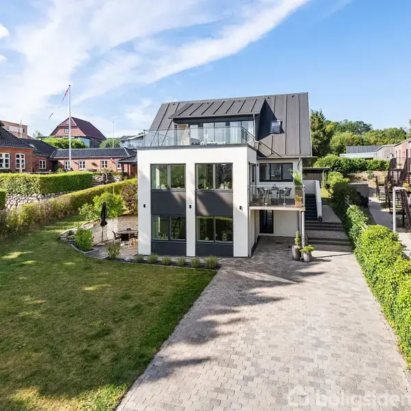 En moderne hvid bygning med store vinduer står i en boligområde. En flisebelagt indkørsel fører op til huset, omgivet af velplejede hække og naboer på begge sider.