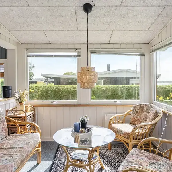 Et lyst rum med rottingmøbler står omkring et rundt glasbord, hvor en lampe hænger over. Stort vindue i baggrunden viser udsigt til have og huse.