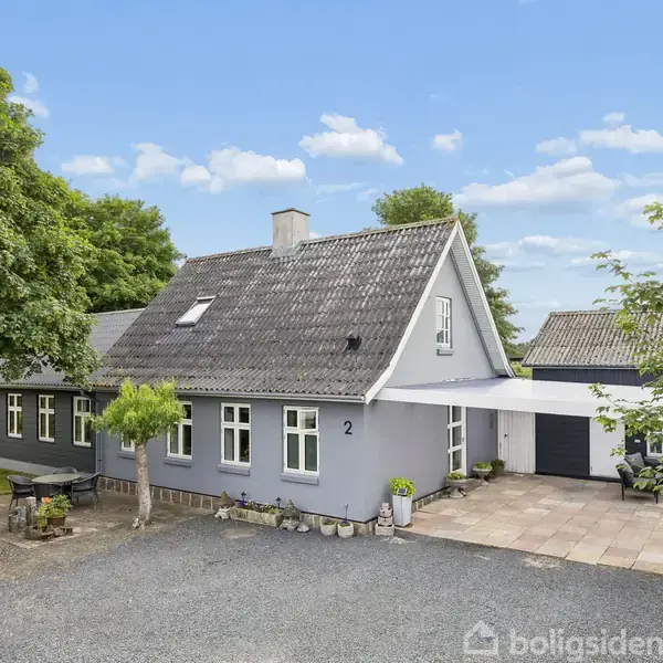 Et gråmalet hus med gråt skråtag står stille omgivet af grønne træer. Adressen "2" ses på facaden. Grusvej og flisebelagt indkørsel leder op til huset.