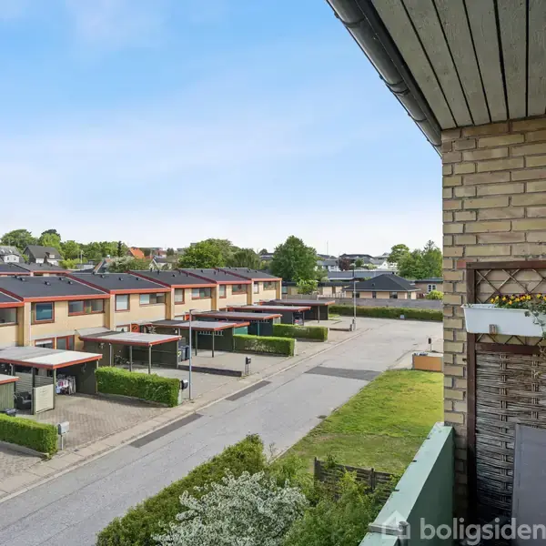 Balkon med plantekasse hænger over en stille vej med rækkehuse. Trælameller på murværk skaber privatliv. Rækkehusene har carporte og små forhaver. Himlen er klar og blå.