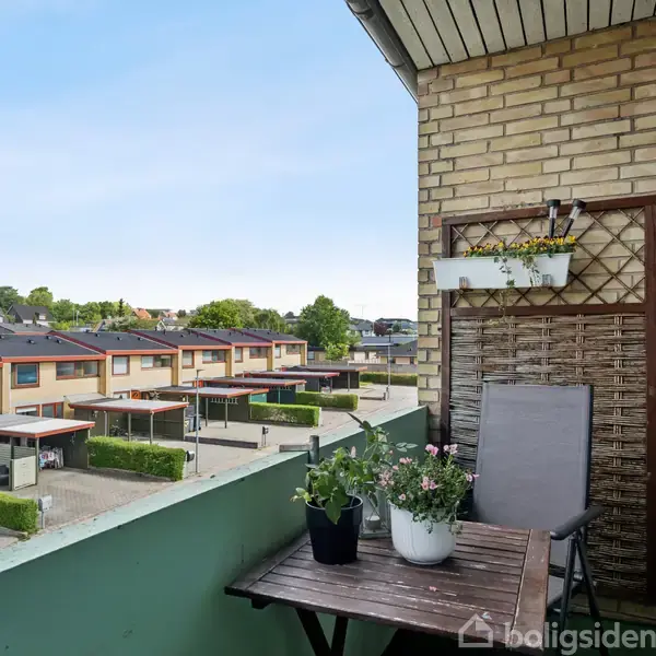 Balkon med træbord og to potteplanter placeret mod venstre. Stol står ved siden af. Udsigt over boligkompleks med rækkehuse i baggrunden under en klar himmel.