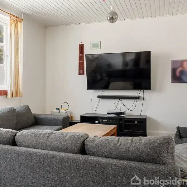 Sofa arrangeret i en U-form står foran fjernsynet på en væg. Rummet har træloft, vindue med gardiner og et sofabord. En grå hundeseng ses i hjørnet.