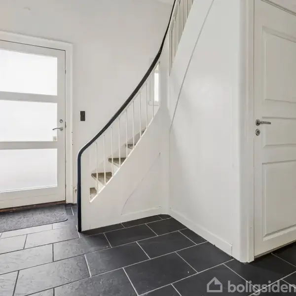 Entré med sort flisegulv og en hvidmalet trappe, der fører opad. Døren har matteret glas og en hvid ramme. Vægge og døre er holdt i hvidt.