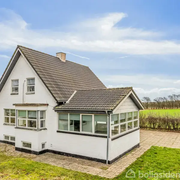 Et hvidt hus med sort tag står stille, omgivet af åbne marker og en flisebelagt terrasse. Ejendommen har en udestue og klare blå himmel i baggrunden.