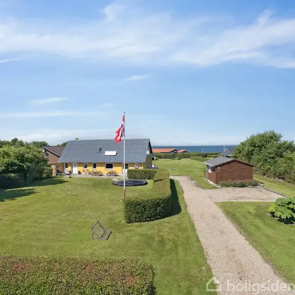 Gult hus med gråt tag står i en stor have. En flagstang med dannebrog er placeret foran huset. Grusvej fører til garage, omgivet af grønne buske og træer.