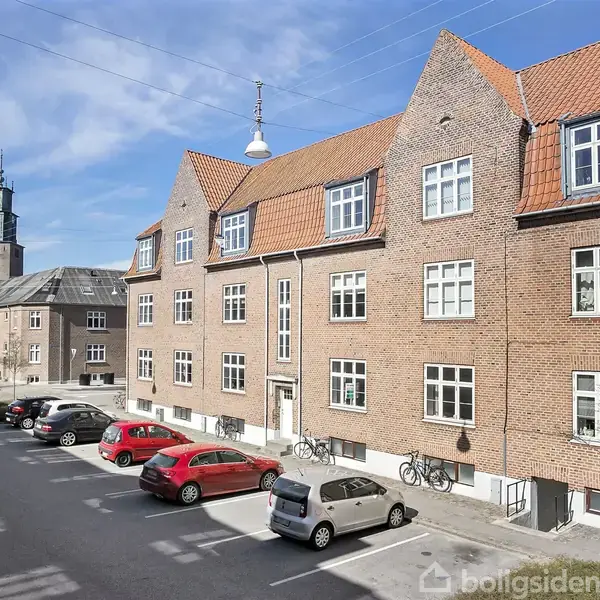 Murstensbygning med rødt tag står langs en vej med parkerede biler og cykler. I baggrunden ses en kirke med et klokketårn under en blå himmel.