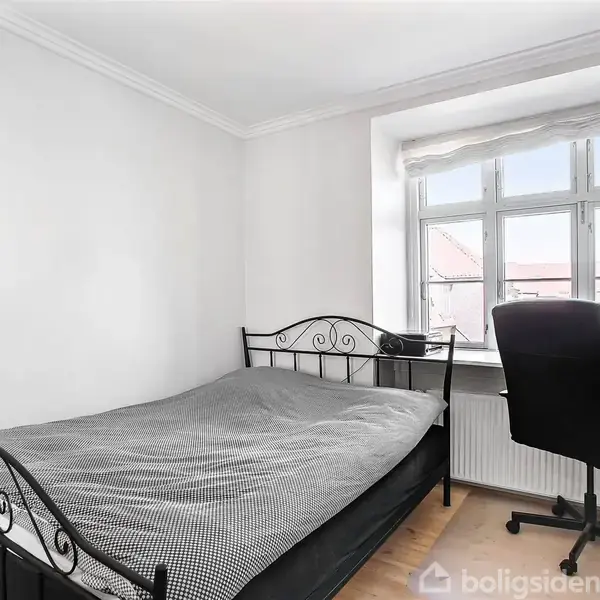 Seng med sort metalramme står i et minimalistisk soveværelse. Ved siden af er et skrivebord med stol foran et vindue, der lukker naturligt lys ind i rummet.
