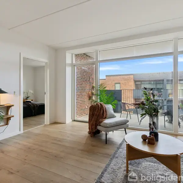 Stue med sofagruppe placeret på et tæppe, sofabord med dekorative genstande, stor glasdør til altan med planter. Hylde med bøger og lampe på væggen til venstre.