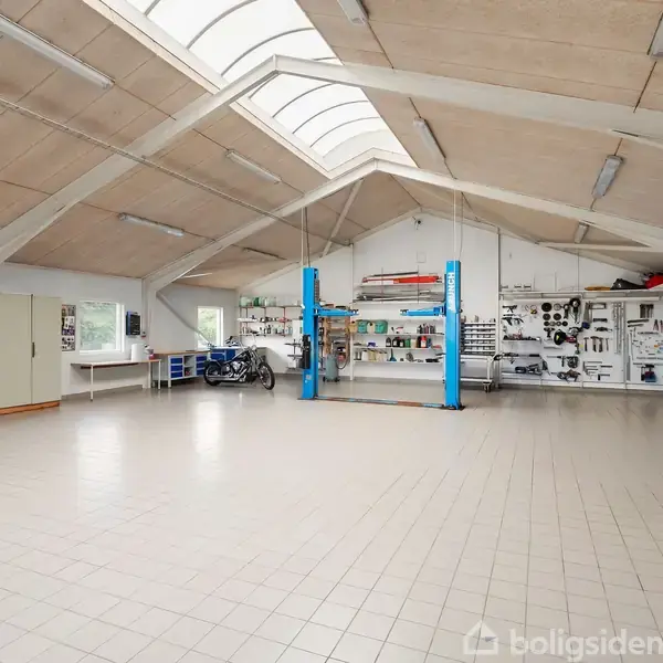 En stor, lys garage med et bil-løfteanlæg centralt placeret. Omkring er der værktøjsreoler og arbejdsborde. Loftet er skråt med ovenlys, og en motorcykel står ved væggen.