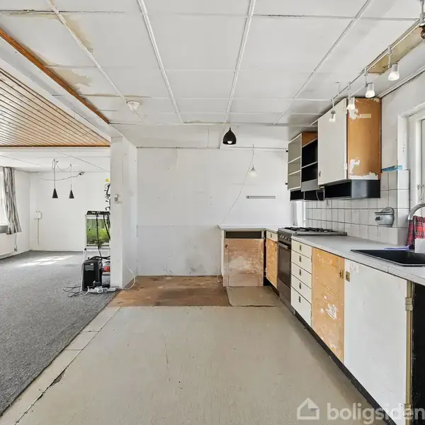Et køkken med åbne hylder og en vask ved vinduet i et lyst rum. Til venstre ses en stue med grå tæppebelægning og store vinduer.