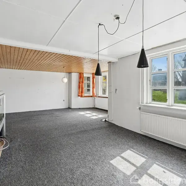 Hvidt hus med skråt tag står ved grusvej omkranset af andre huse. Sollys kaster skygger, og en plæne løber langs siden.