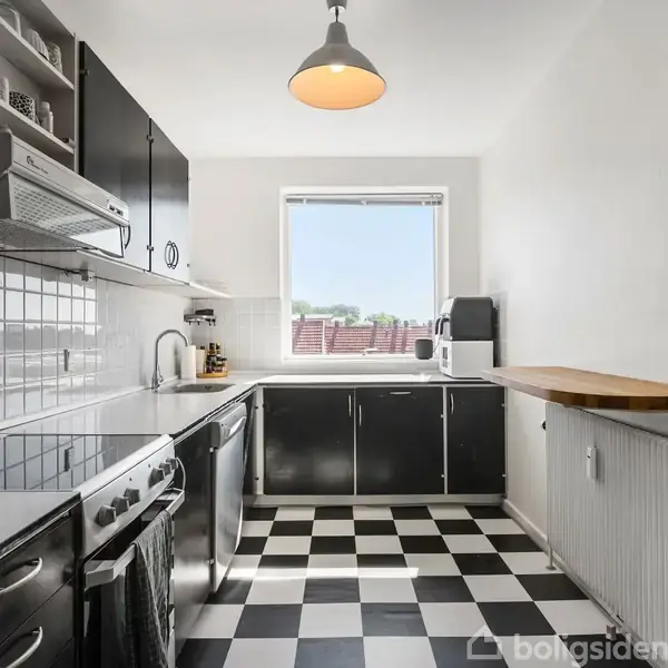 Et køkken med sorte og hvide ternede fliser på gulvet. Skabene er sorte, der er hvide fliser på væggen, en ovn til venstre og et vindue med udsigt. En træbordplade ligger over en radiator til højre.