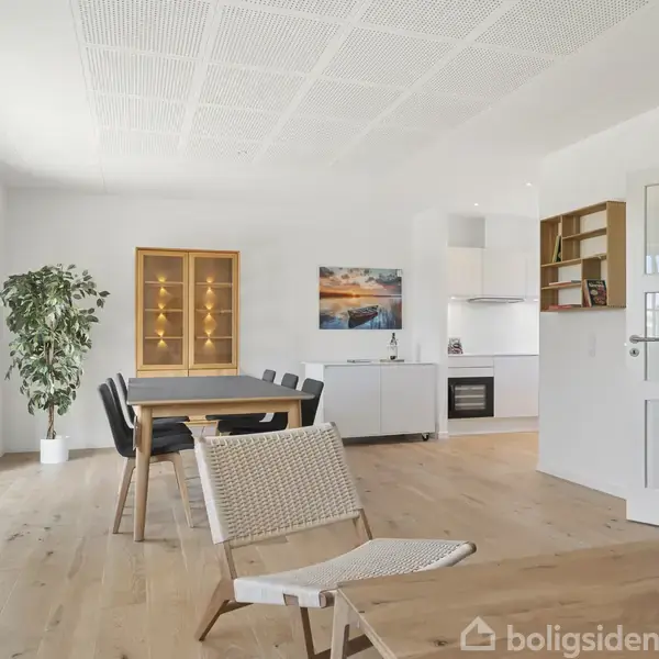 En moderne stue med trægulv, indeholder et spisebord, stole og en lænestol. Et glasskab og en plante står i venstre side. Køkkenet ligger bag ved spiseområdet.