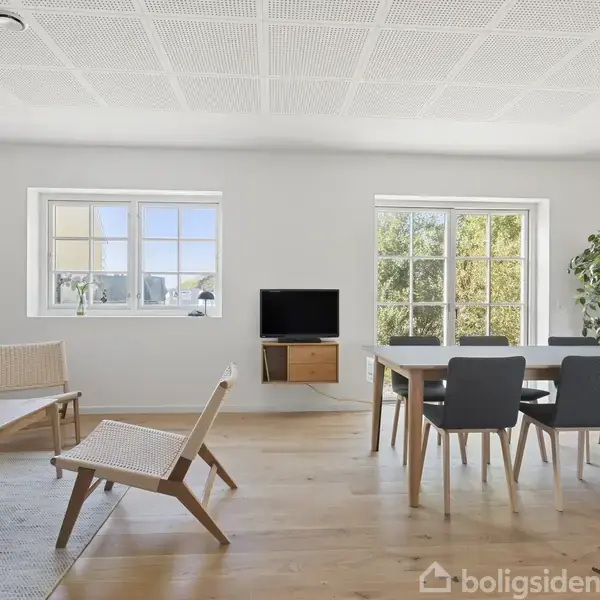 Stue med træmøbler, sofabord og stole omkring spisebord. Fjernsyn på væg og stor plante ved vindue. Lyse vægge og trægulv skaber et lyst miljø.