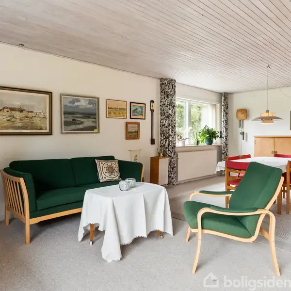En stue med en grøn sofa og stol arrangeret omkring et bord med en hvid dug, omgivet af kunst på væggene og et spisebord ved vinduet.