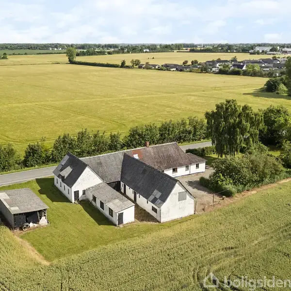 Et hvidt landhus med gråt tag står på en grøn mark ved en vej. I baggrunden findes træer og en landsby.