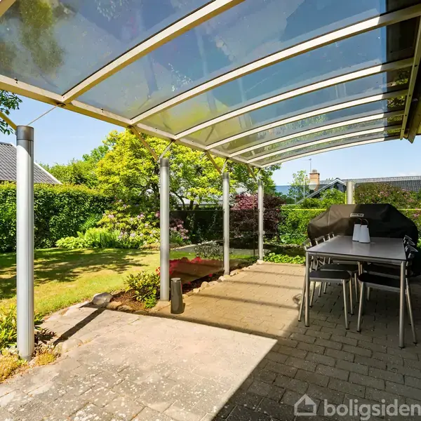 Terrasse med glasoverdækning og spisebord, der står stille. Omgivet af en frodig have med buske og træer under en klar blå himmel.