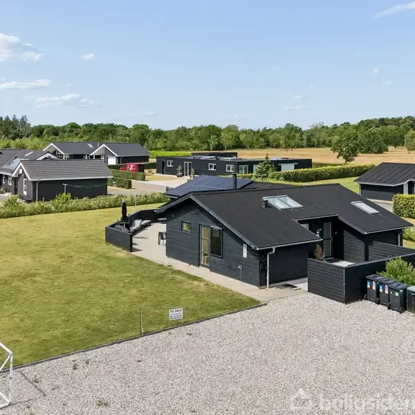 En sortmalet enfamilievilla med tagvinduer står stille på en grusindkørsel. I baggrunden ses en stor græsplæne omkranset af flere lignende huse og skov.