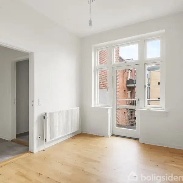 Et lyst værelse med trægulv og hvide vægge åbner sig mod en balkon gennem store vinduer. En åben dør fører til et tilstødende, flisebelagt rum.