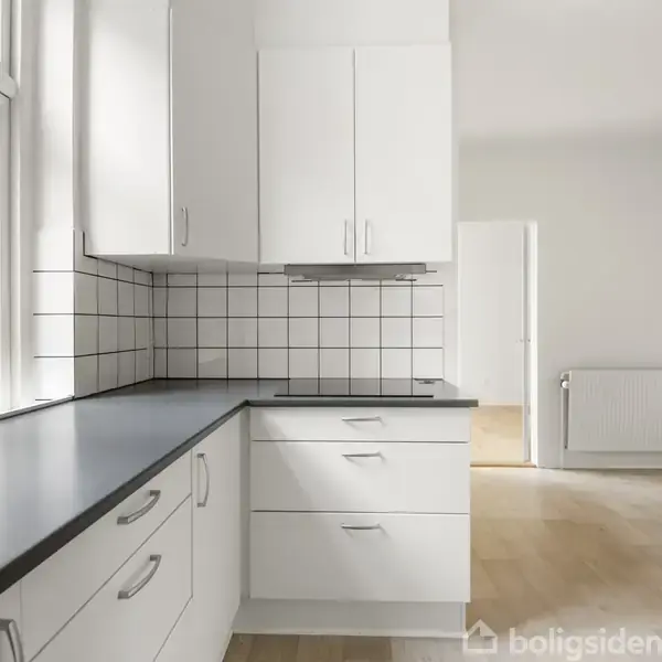 Køkken med hvide skabe og sorte bordplader, arrangeret i hjørne. Flisebelagt væg ved komfur. Ved siden af er en lys stue med trægulve og radiator.