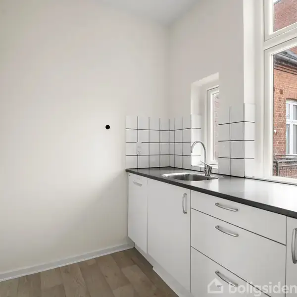 Hvide køkkenskabe står langs væggen med en indbygget vask og flisevæg. Store vinduer åbner op til udsigt mod rødt murstensbyggeri. Trægulv løber gennem rummet.