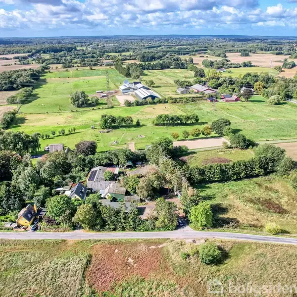 Landskab med gårde og huse omgivet af grønne marker og skove. En vej løber langs forgrunden, og spredte skyer fylder den blå himmel.