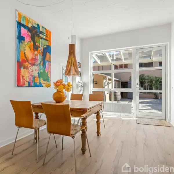 Træbord omgivet af fire stole med en vase fyldt med blomster står i et lyst rum. Store malerier hænger på væggene, og en glasdør leder ud til en terrasse.