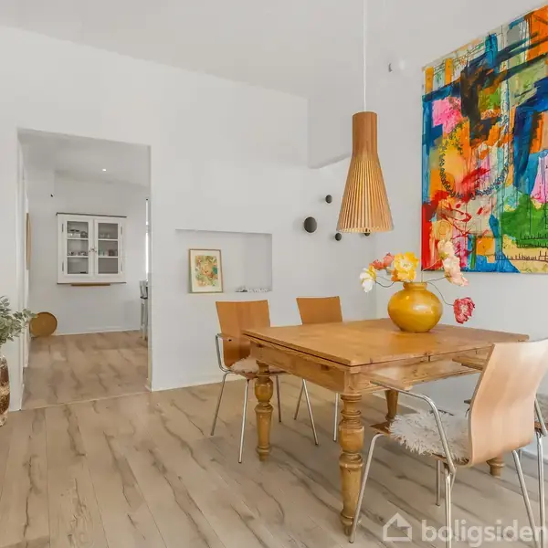 Et træbord omgivet af moderne stole står i en spisestue med lyst trægulv. Over bordet hænger en moderne lampe, og en farverig abstrakt kunstplakat pryder væggen.